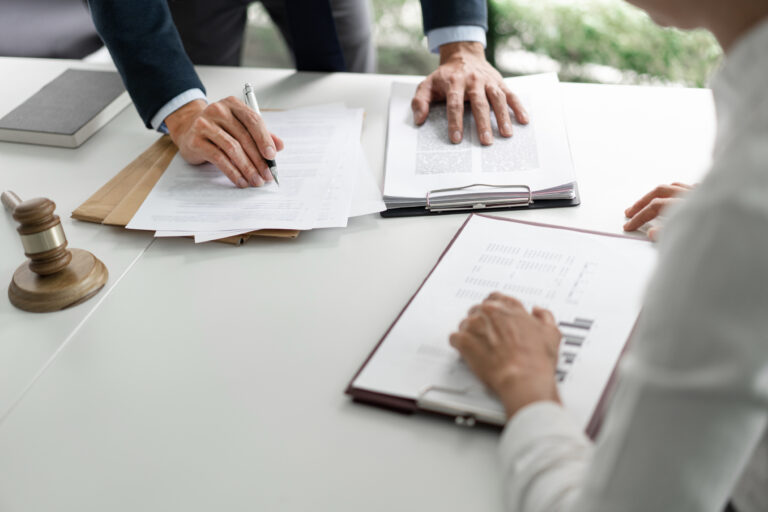 Two people sign and review legal documents on a white desk, with a gavel and envelopes nearby.