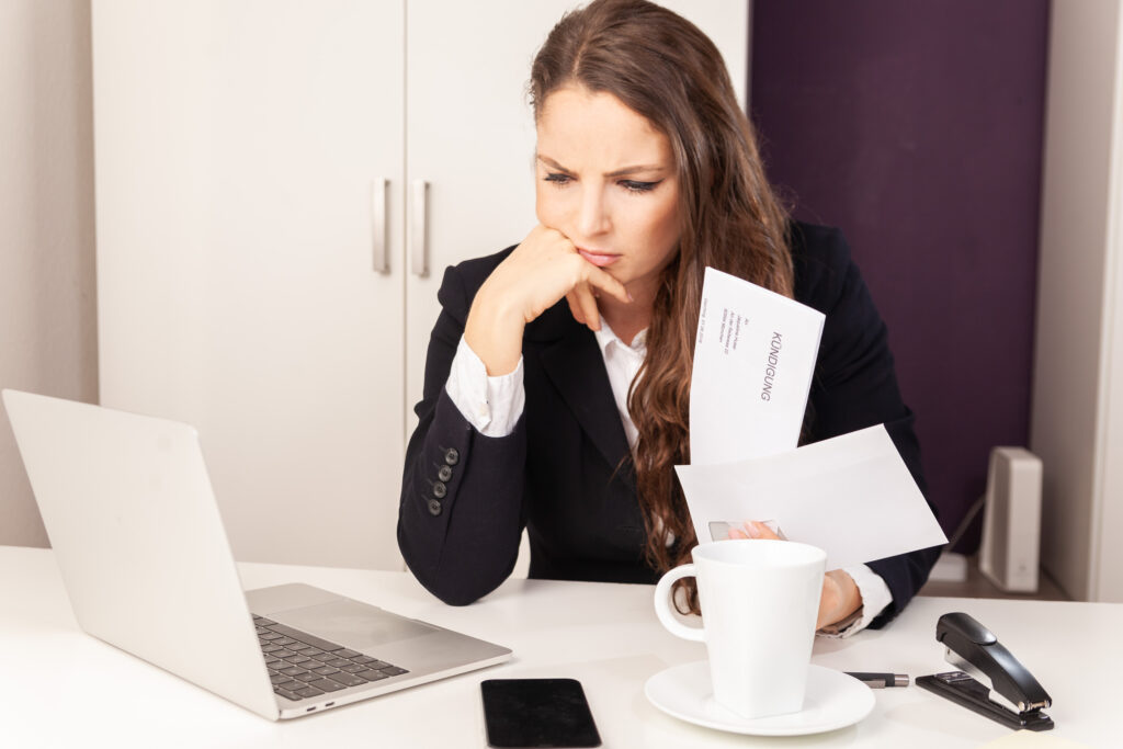 Businesswoman in a black blazer sits at a white desk, frowning at papers while looking at a laptop nearby.